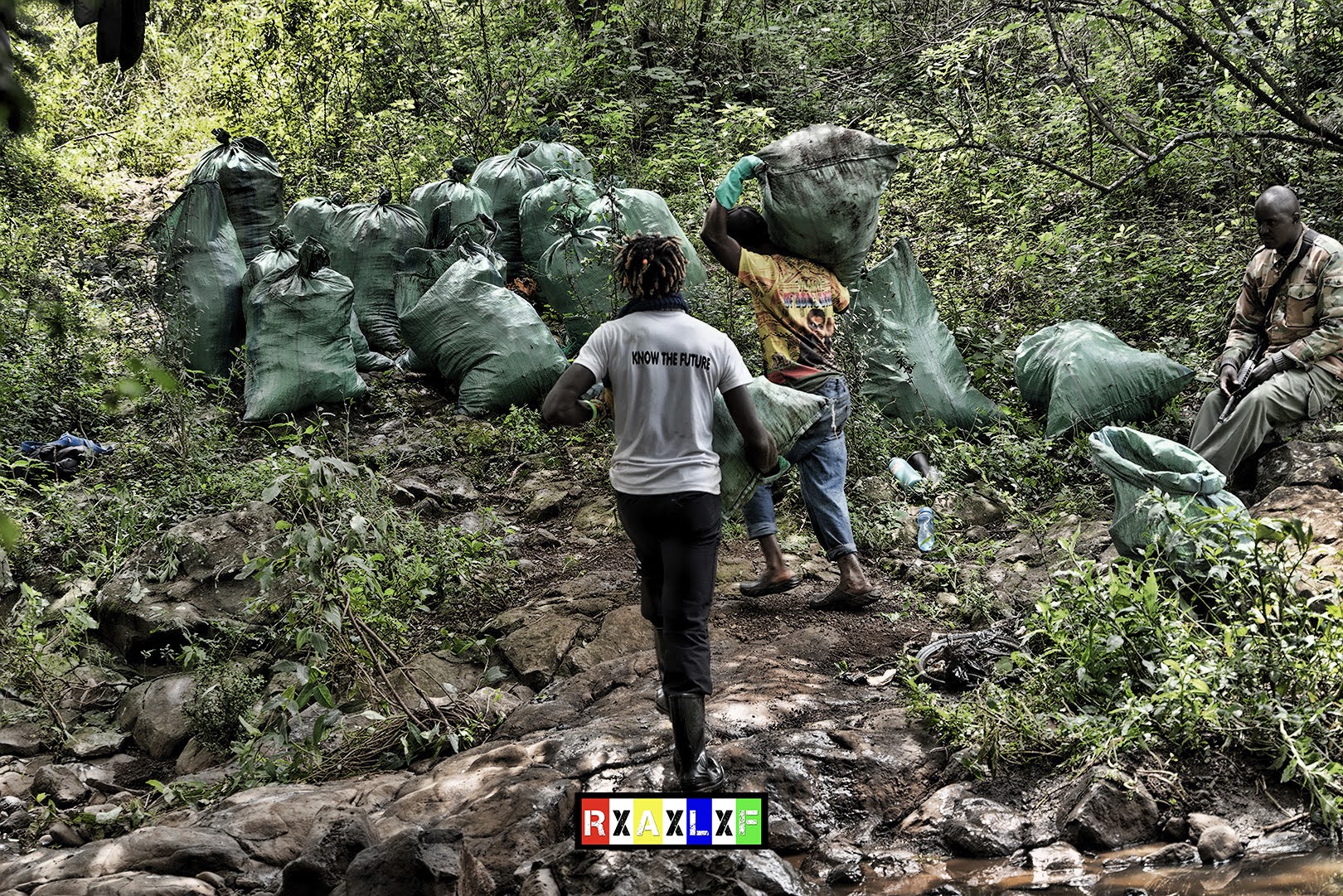 Two volunteers carrying large green sacks of collected trash through a forested area, with more filled sacks piled in the background. A ranger sits nearby, overseeing the cleanup efforts. The scene captures the dedication and teamwork involved in the River Kandisi cleanup.