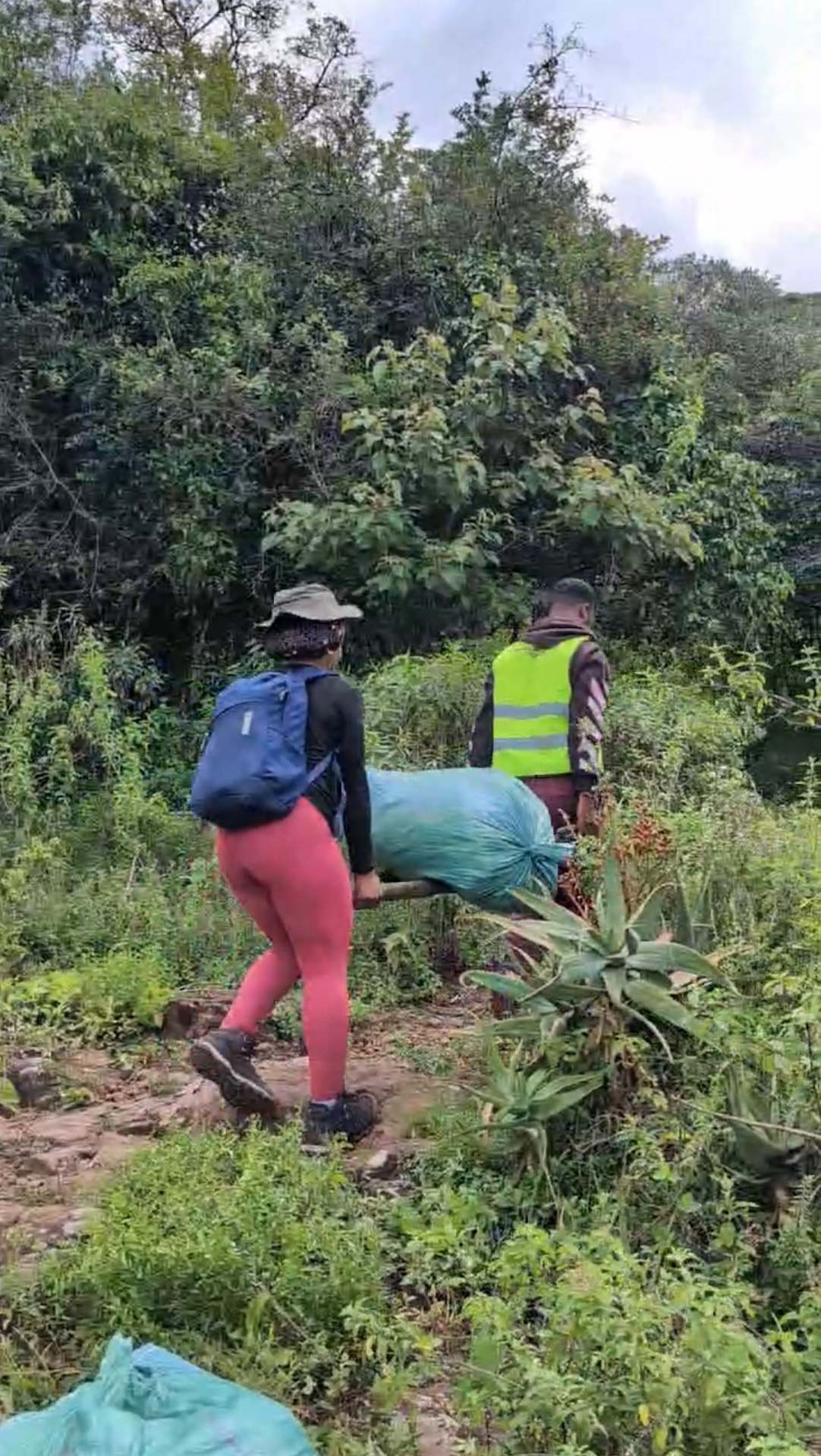 Two volunteers, one wearing a blue backpack and another in a reflective vest, carrying a large green bag of collected waste along a path surrounded by dense greenery during the River Kandisi cleanup